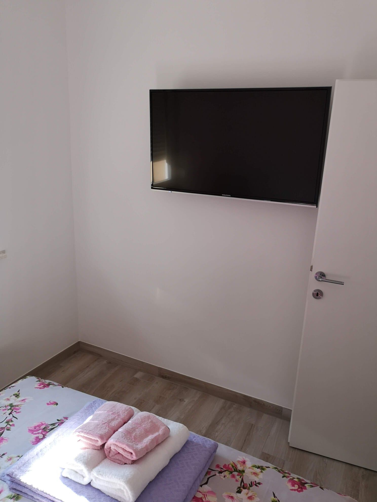 A small room with a bed, a flat-screen TV mounted on the wall, and a door.