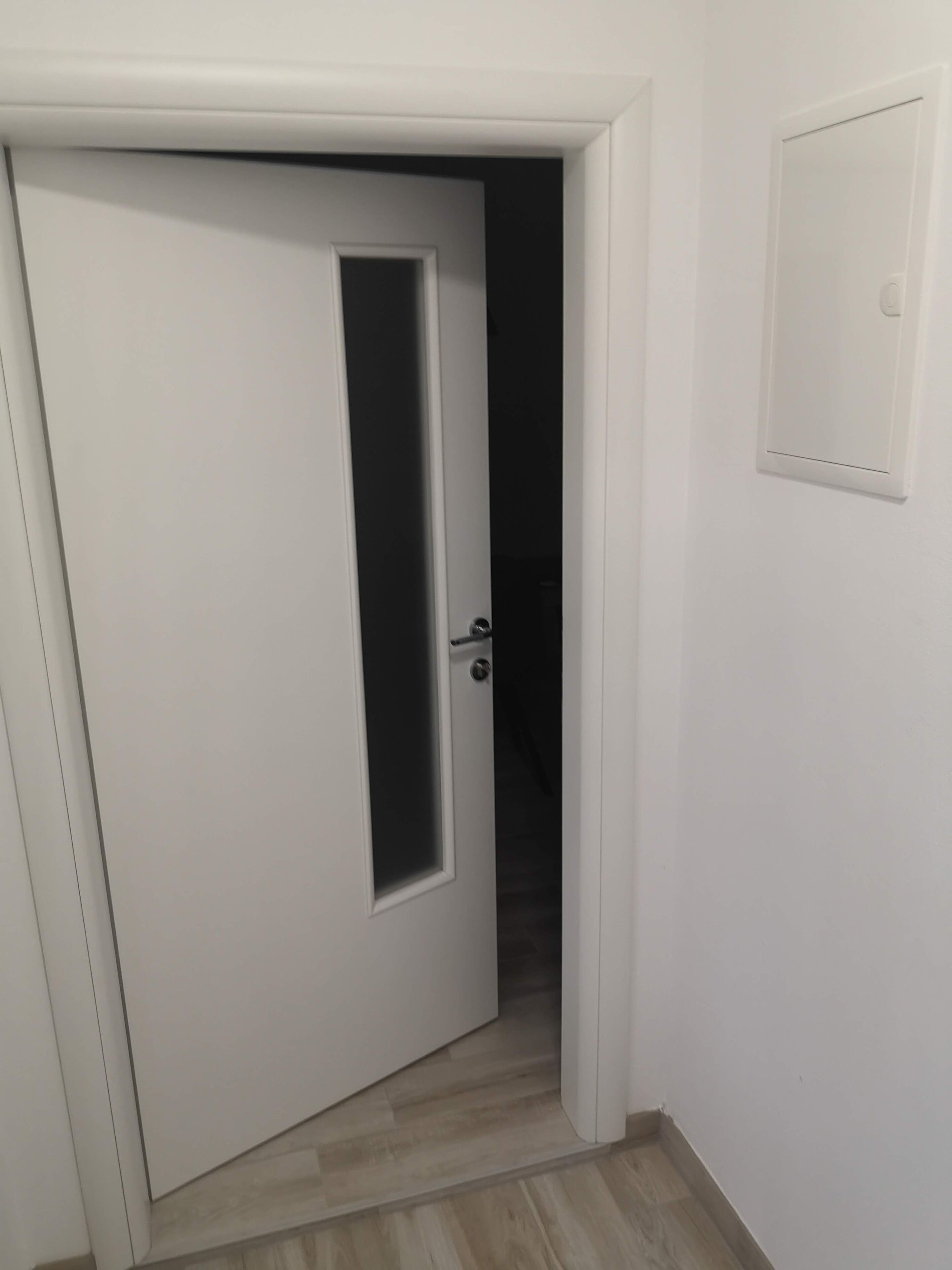 A white door with a small window, a light switch on the wall, and a wooden floor.