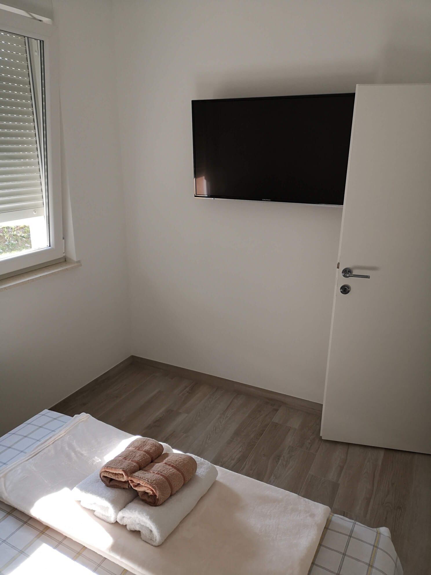 A room with a bed, a flat-screen TV mounted on the wall, a door, and a window with blinds.