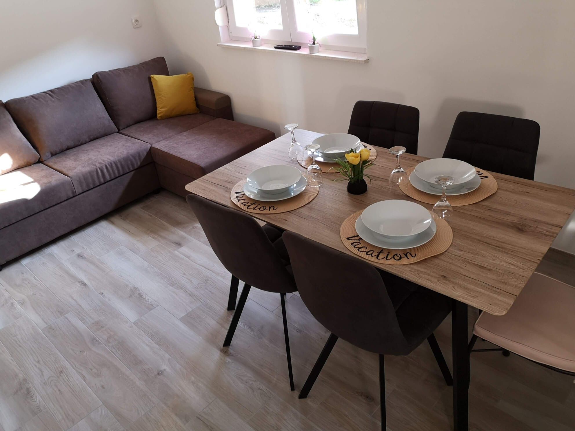 A dining area with a wooden table set for four, surrounded by dark chairs. A brown sofa with a yellow pillow is against the wall, and a small window with a plant is visible.