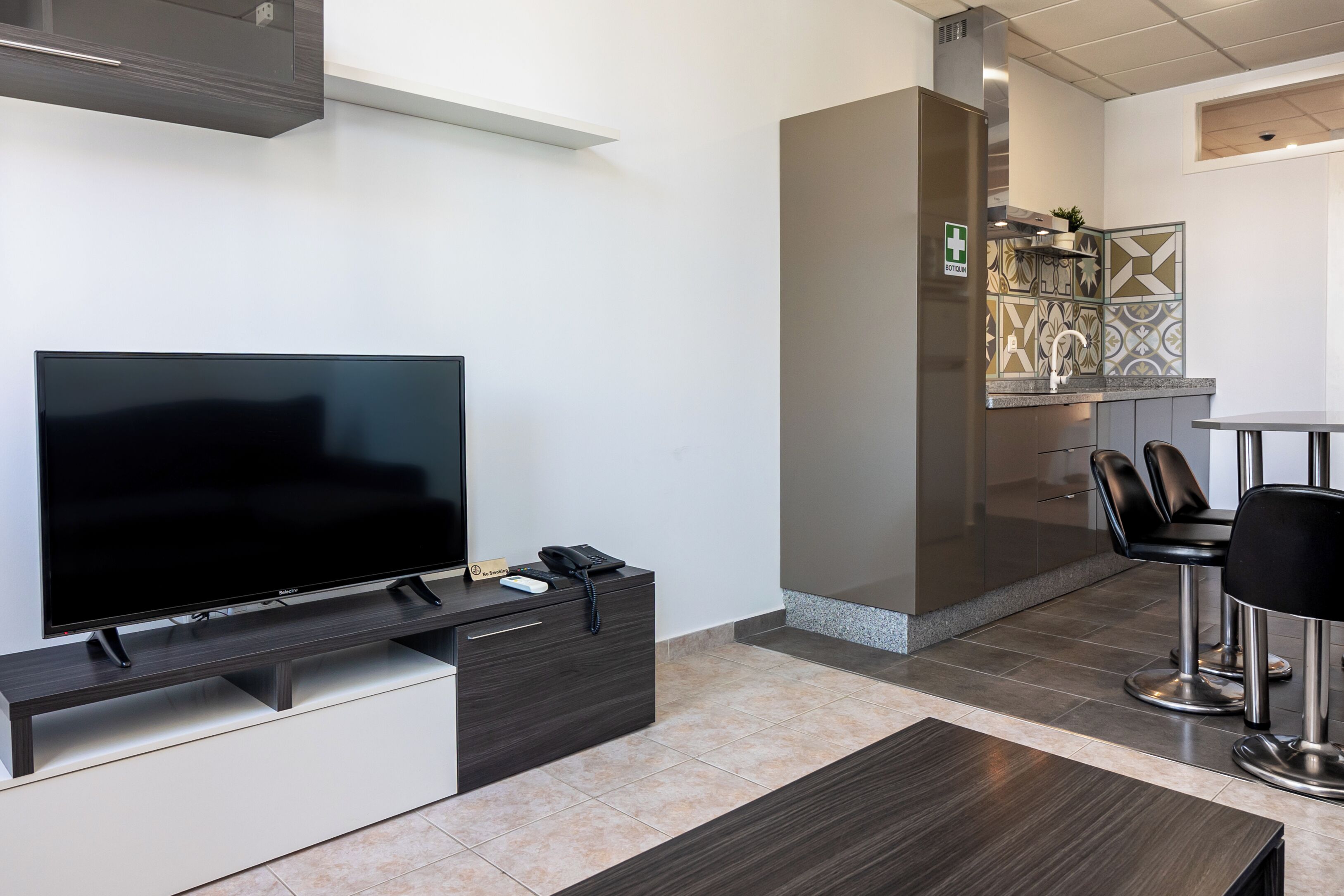 Ein modernes Wohnzimmer mit Flachbildfernseher, einer dunkelholzartigen Ablage, einem Essbereich mit Tisch und Stühlen sowie einer Küche mit dekorativer Rückwand.