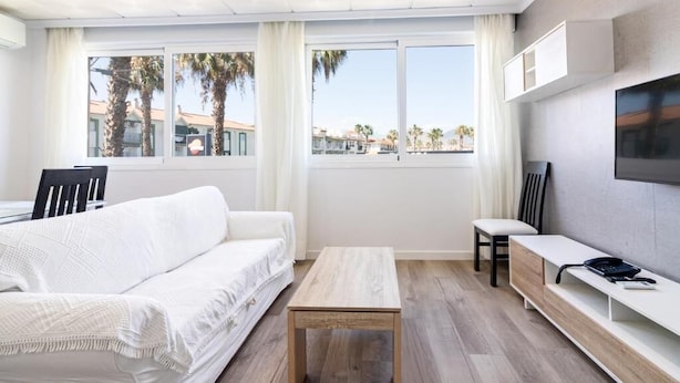 Una sala de estar moderna con un sofá blanco, una mesa de centro de madera y un televisor montado en la pared. Hay dos ventanas con cortinas que ofrecen vistas a palmeras y edificios.