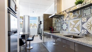 Cocina moderna con refrigerador de acero inoxidable, estufa integrada y salpicadero decorativo. Se ve un ventanal con vista a palmeras y un comedor con dos sillas.