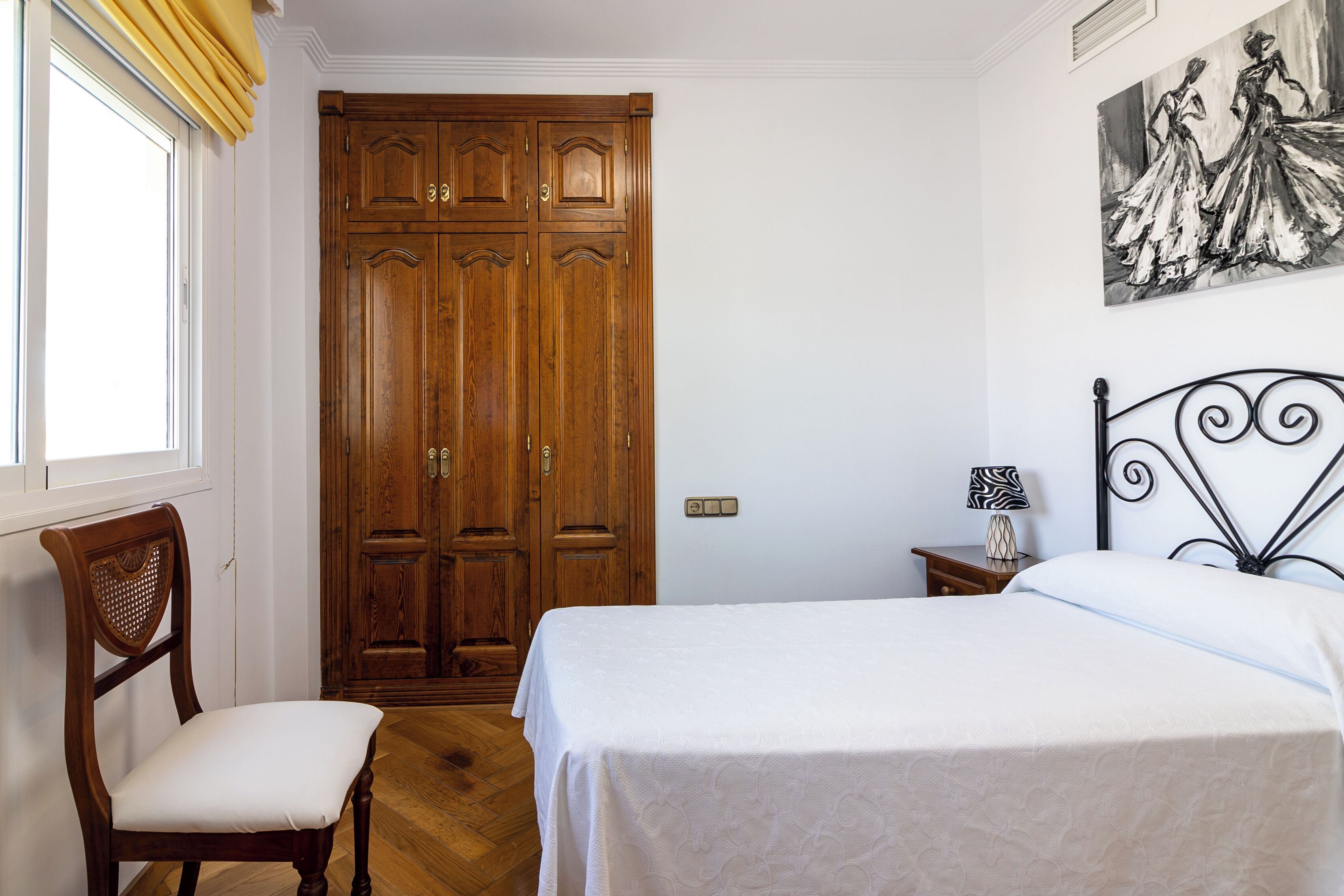 Un dormitorio con un armario de madera, una cama con cubrecama blanco, una silla, una mesita de noche con lámpara y un cuadro en la pared.