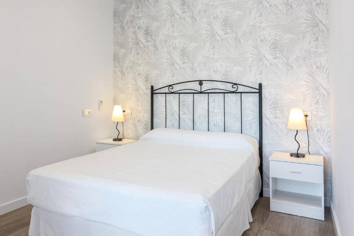 Un lit simple avec une tête de lit en métal, deux tables de chevet avec des lampes et un papier peint à motifs sur le mur.