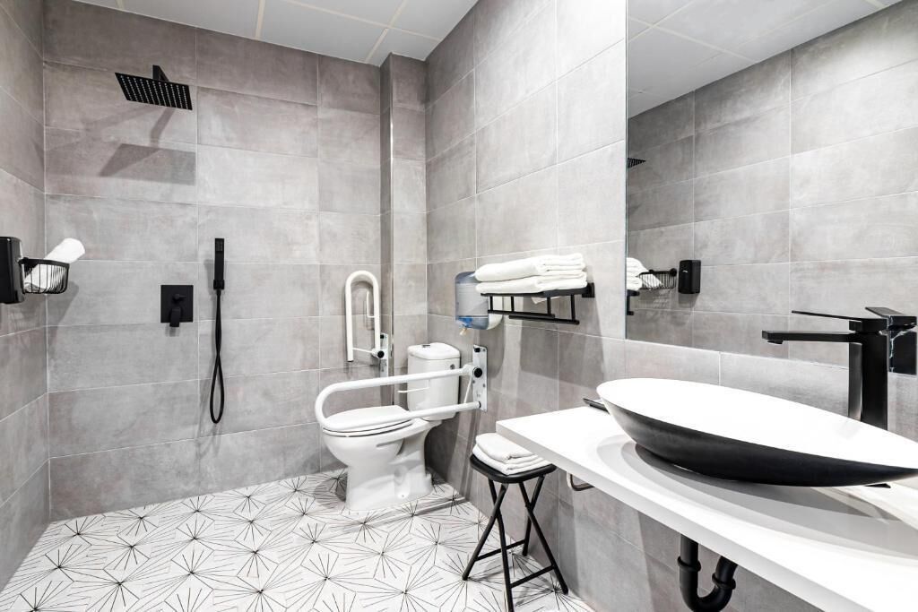 Une salle de bain moderne dotée d’une grande baignoire, de toilettes équipées d’une barre d’appui, d’une pomme de douche et d’un lavabo avec un grand bac.