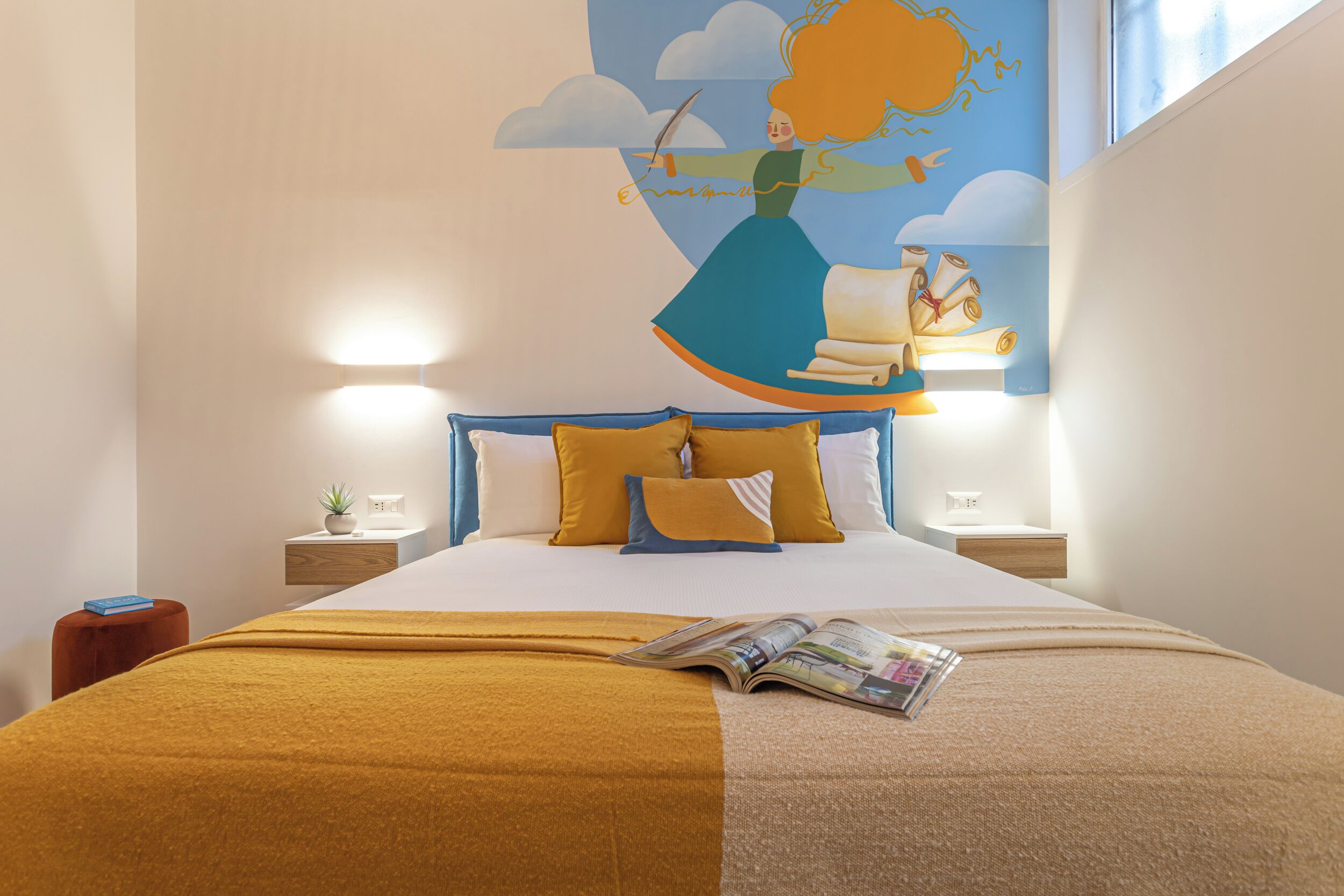 A neatly made bed with a magazine, a blue headboard, and a wall art depicting a woman flying.