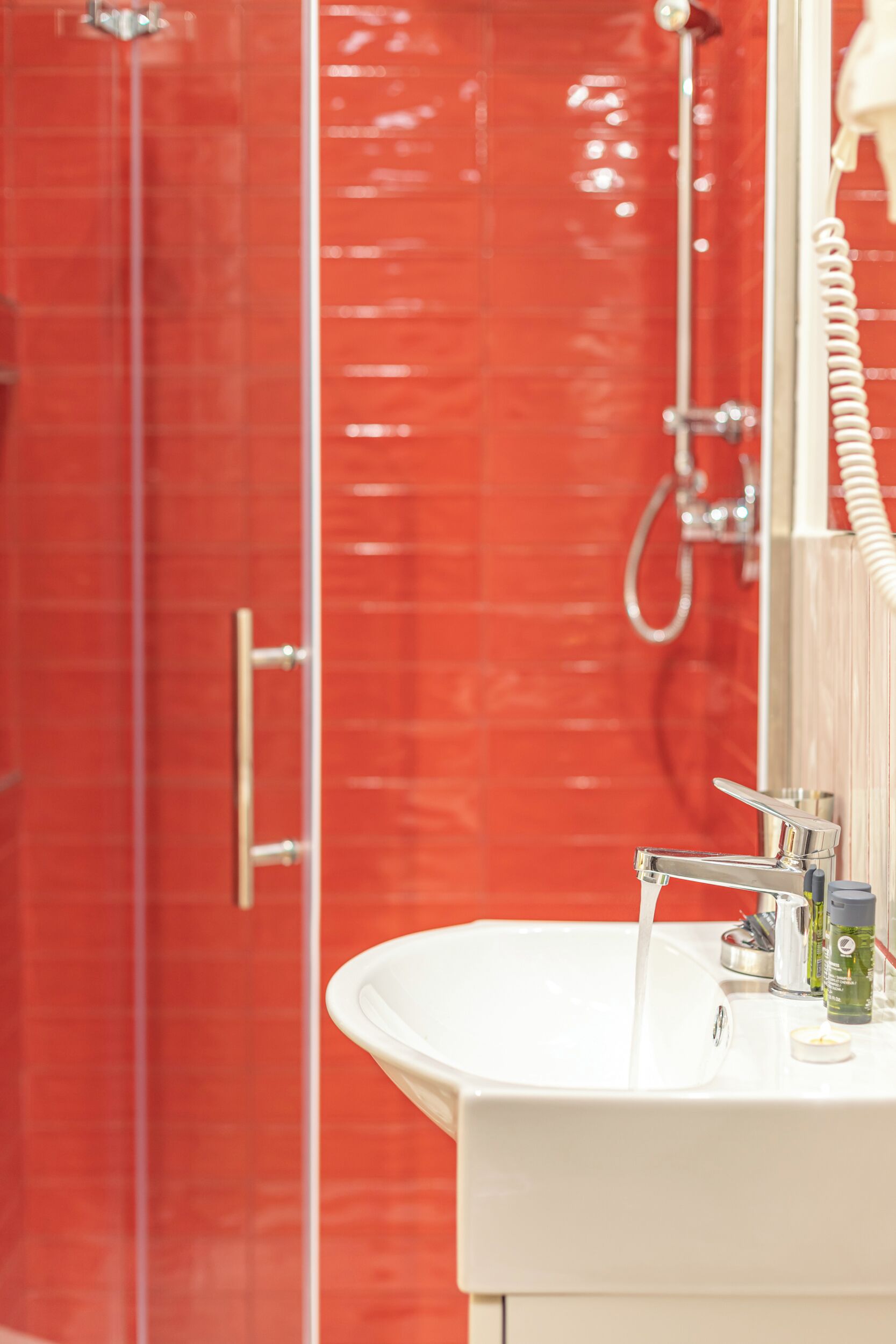 A bathroom with a red tiled wall, a white sink with running water, and a handheld showerhead.