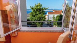 A balcony with a view of a tree and buildings, a chair, and a window with curtains. - Apartman Mira (Crikvenica)