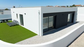 Ein modernes Gebäude mit flachem Dach, einer kleinen Terrasse mit Kunstrasen und Liegestühlen sowie einem Kiesweg, der zum Eingang führt.
