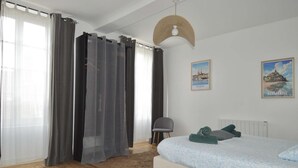 Un dormitorio con una cama, una silla, un ventanal con cortinas y dos cuadros colgados en la pared.