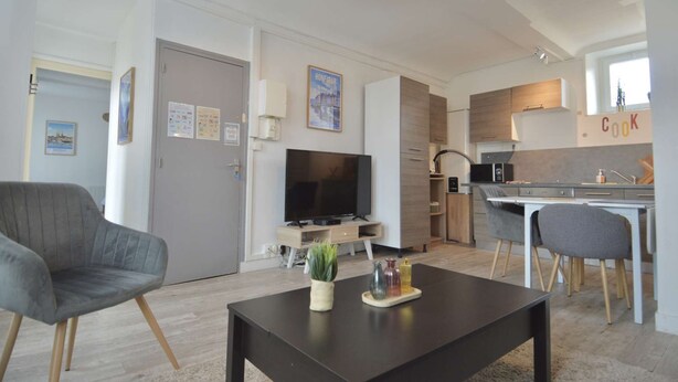 Una sala moderna con un televisor de pantalla plana, una mesa de centro con objetos decorativos, un sillón gris y una zona de comedor con una cocina al fondo.
