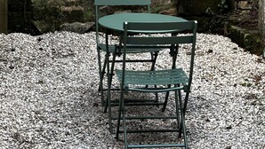 Una mesa y sillas sobre grava, con un muro de piedra al fondo.