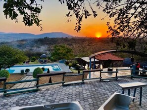 A pool area with hexagonal tiles, a gazebo, and a view of the sunset over a hilly landscape. - The Rock at Edwaleni (Matsapha)