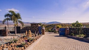 A paved entrance with a stone wall, palm trees, and a view of distant mountains. - The Rock at Edwaleni (Matsapha)