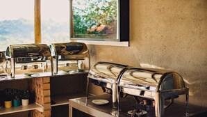 A breakfast station with two stainless steel coffee dispensers, a brick counter, and a window with a view of greenery. - The Rock at Edwaleni (Matsapha)