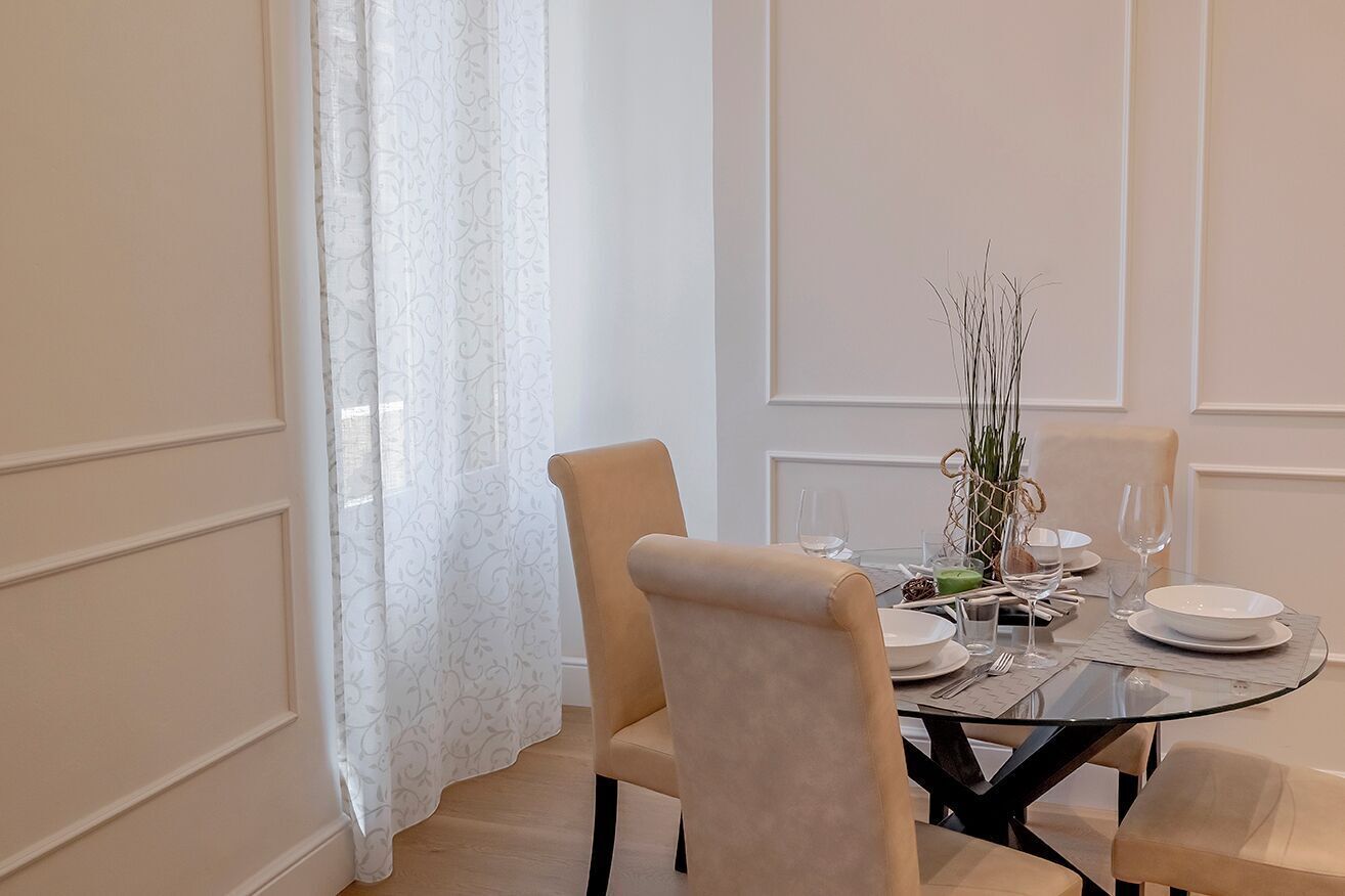 Zona pranzo con tavolo rotondo in vetro, sedie imbottite color beige e tovaglia bianca.