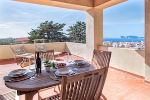 A wooden table set for dining with plates, glasses, and a bottle on a balcony overlooking a coastal town. - Sardinia RE - Dimora Della Contessa (Alghero)