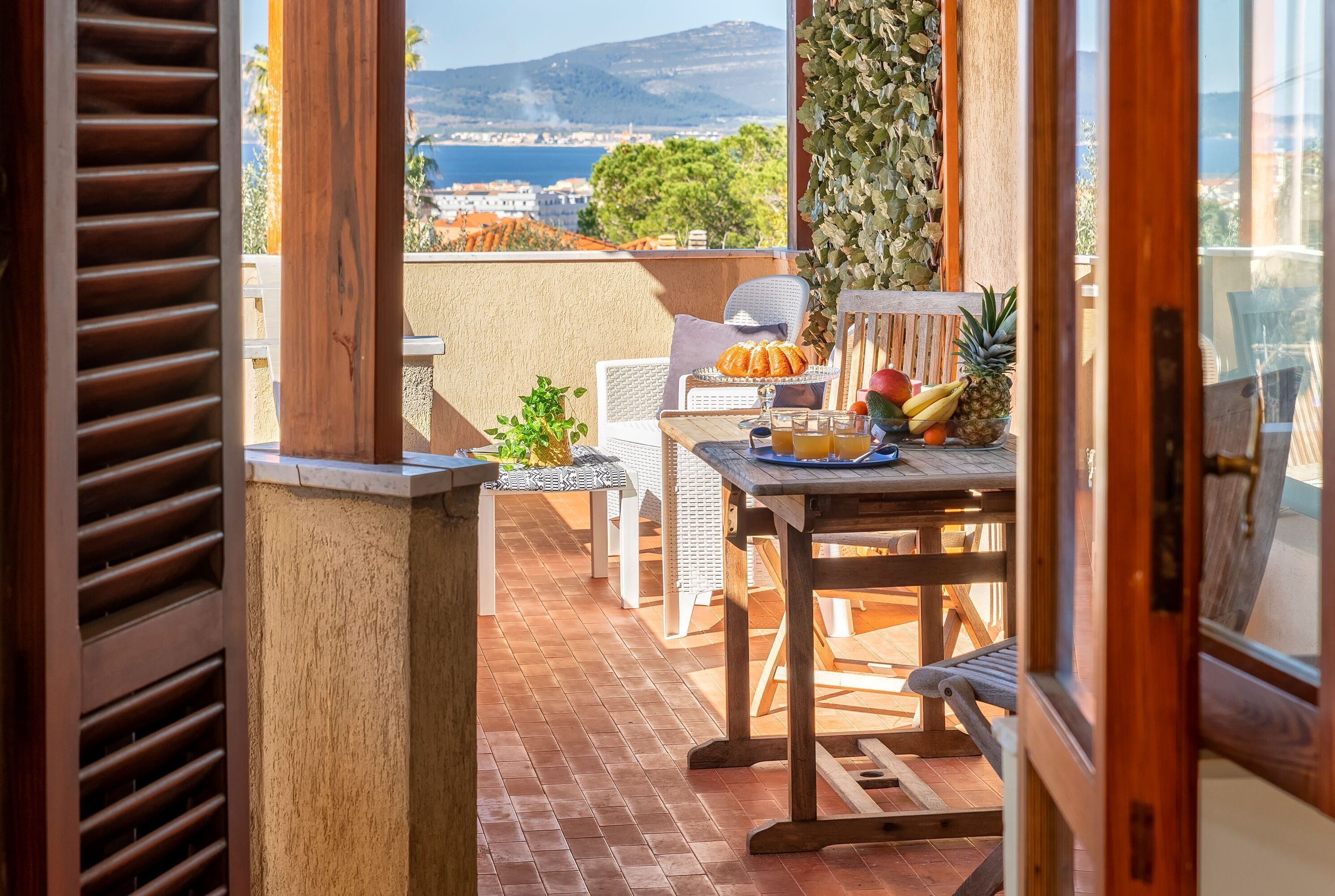 A balcony with a table set for dining, featuring bread, fruits, and a glass of drink.