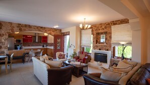 A spacious living area with a stone wall, a fireplace, and a kitchen in the background. - Villa Angels in Crete (Malevizi)