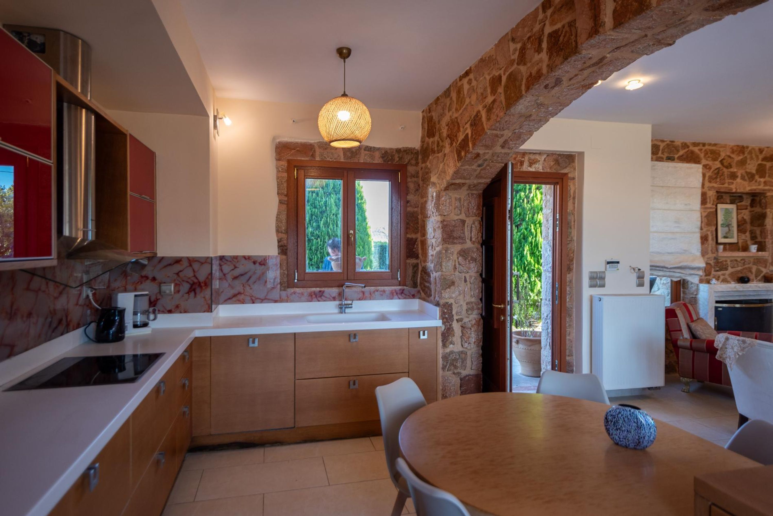 A modern kitchen with a stone archway, a dining area with a round table, and a view of greenery outside.
