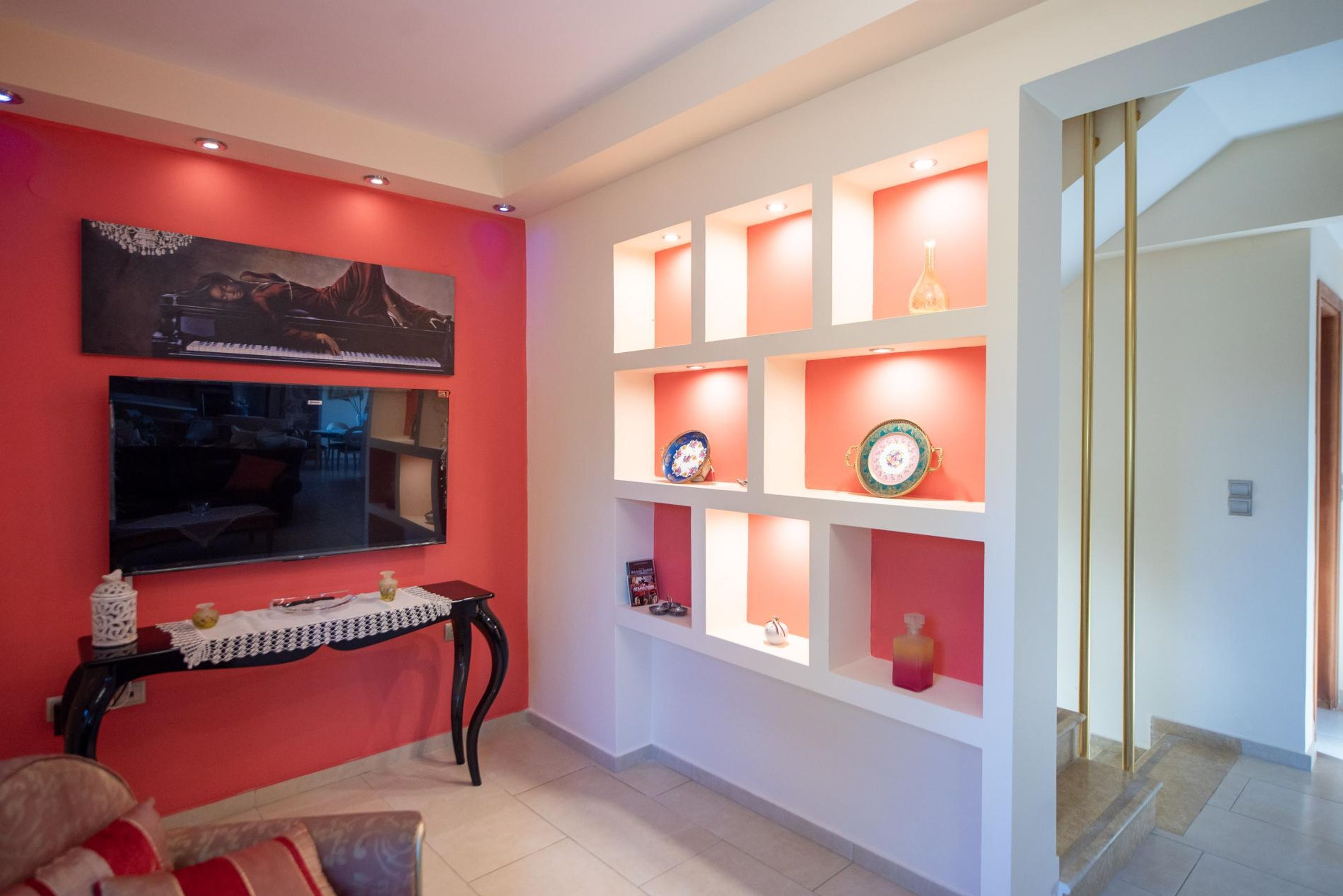 Un salon avec un mur d’accent rouge, un téléviseur fixé au mur, une table basse agrémentée d’objets décoratifs et une étagère blanche avec des portes en verre, où sont exposés divers objets.