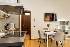 Una cocina moderna con estufa integrada, microondas y cafetera. Hay un comedor con una mesa y sillas blancas, y un televisor en la pared mostrando el logotipo de Netflix.