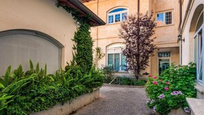 A narrow cobblestone pathway flanked by lush greenery and flowering plants, leading to a building with arched windows and a door. - Colosseum Sweet Life Apartment  (Rome)