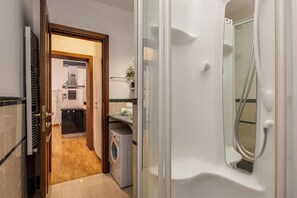 Un baño de hotel con ducha de vidrio, lavadora y un lavamanos pequeño.