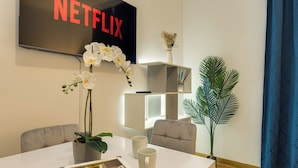 A modern living room with a flat-screen TV displaying 'NETFLIX', a white coffee table with a magazine, two white mugs, a potted orchid, and a tall plant. - Colosseum Sweet Life Apartment  (Rome)