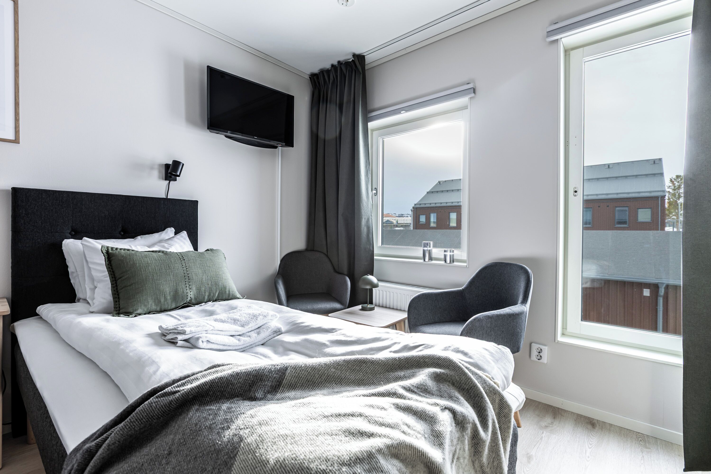 A modern hotel room with a large bed, a television, a chair, and a window with a view of buildings.