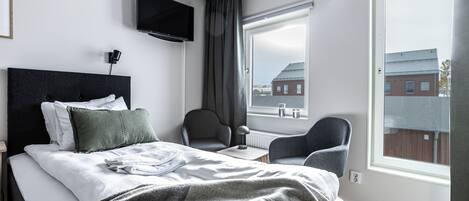 A modern hotel room with a large bed, a television, a chair, and a window with a view of buildings.