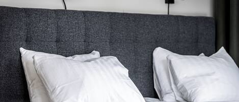 A neatly made bed with white linens and pillows, a grey upholstered headboard, and two wall-mounted black speakers.