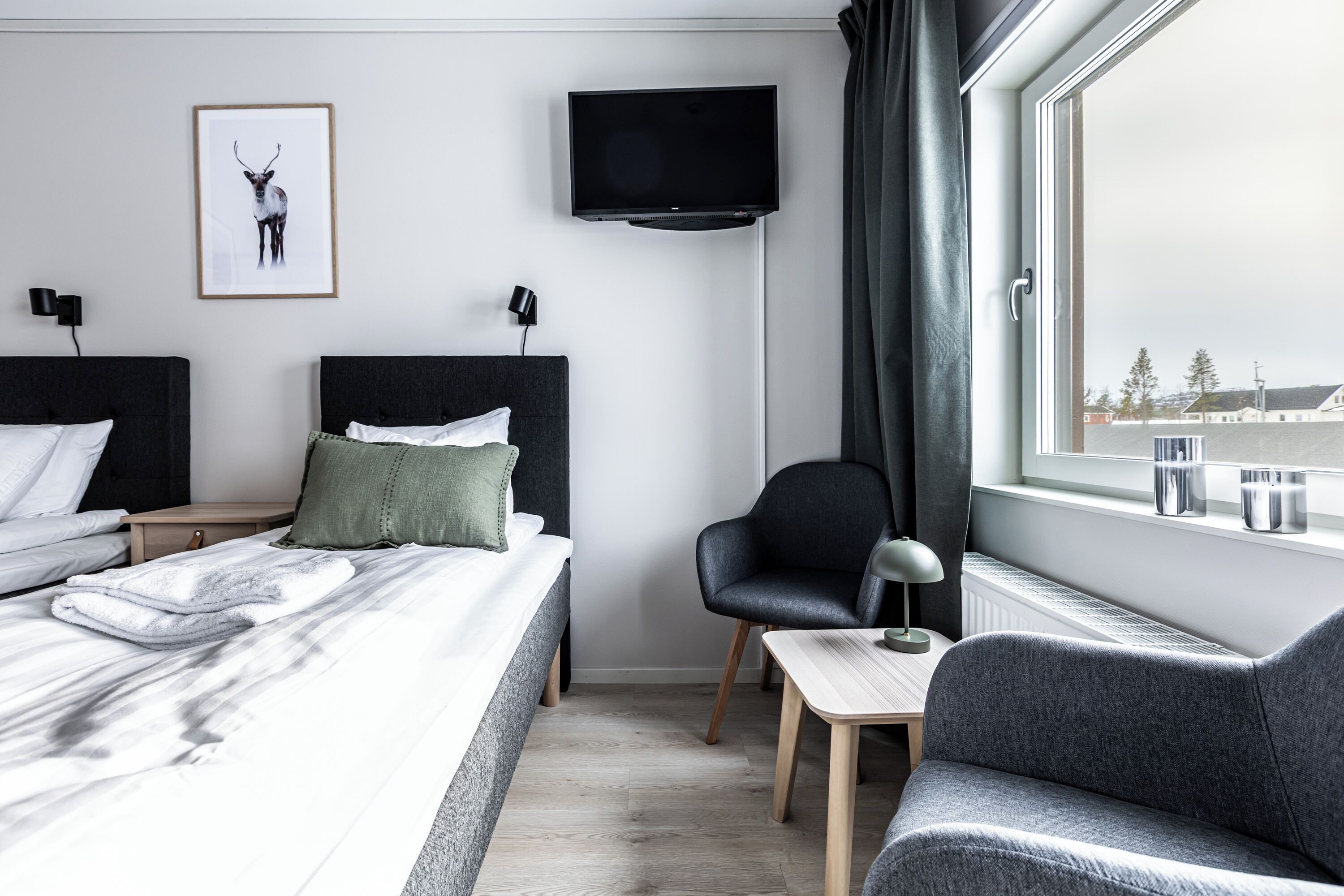 A modern hotel room with a bed, two chairs, a desk, a TV, and a window with a view of buildings.