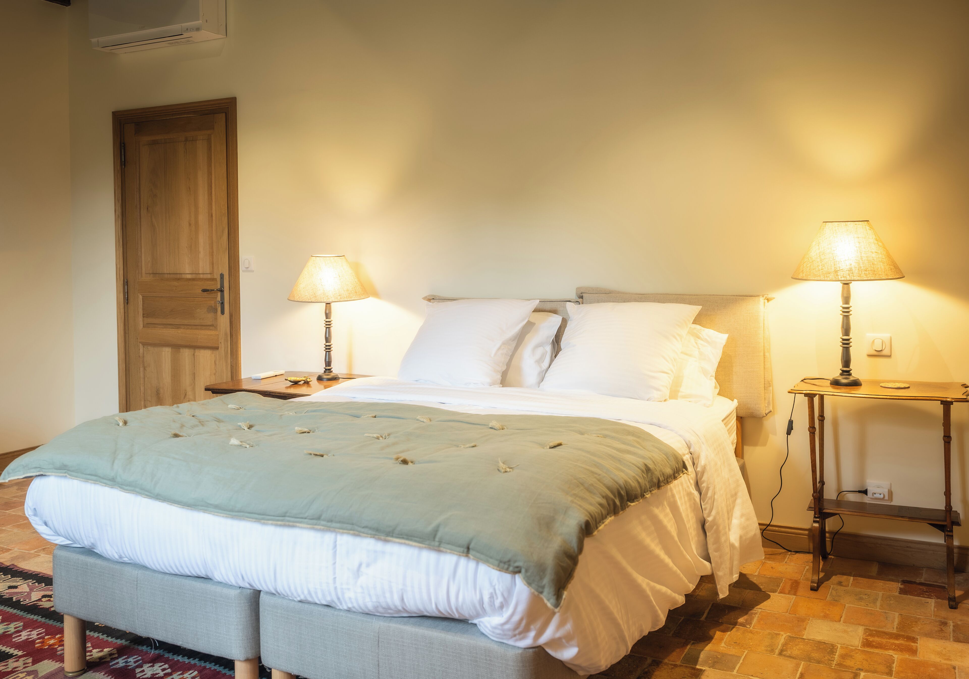 A neatly made bed with a light blue bedspread, two bedside lamps, and a wooden door in a room with a tiled floor.