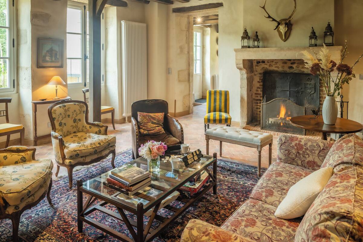 A cozy living room with a fireplace, floral armchairs, a sofa, and a coffee table with books and flowers.