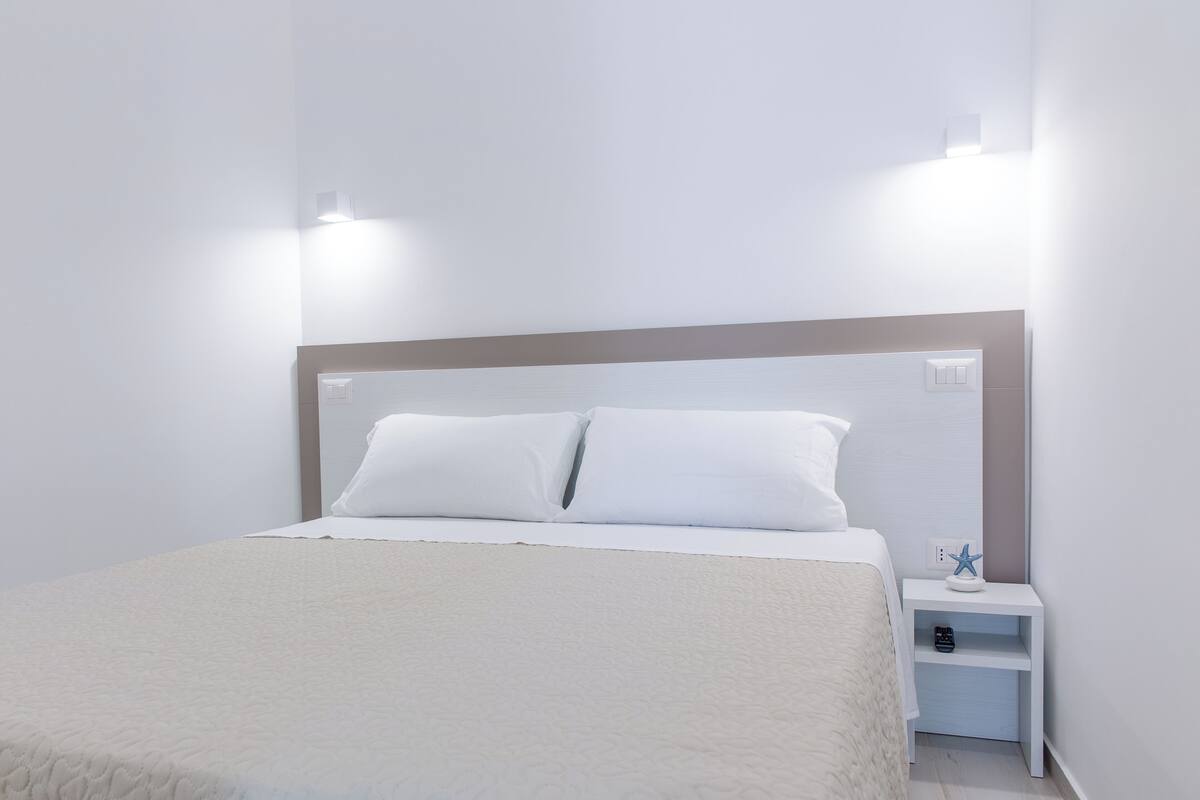 Una cama bien hecha con ropa de cama blanca y un cabecero, una mesita de noche con una lámpara, y un pequeño estante decorado con una estrella.