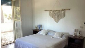 A bedroom with a bed, a nightstand, a wall-mounted macrame piece, and a door leading to an outdoor area. - The house in the woods (Gorgofreddo)