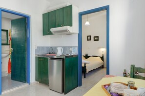 A compact kitchen with a refrigerator, a coffee maker, and a microwave. There is a bed with a white mattress and a blue headboard.
