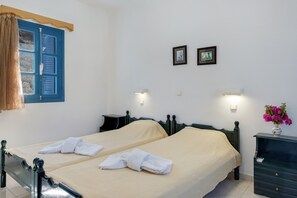 A hotel room with two beds, a window with blue shutters, a nightstand with a lamp, and a vase with flowers.