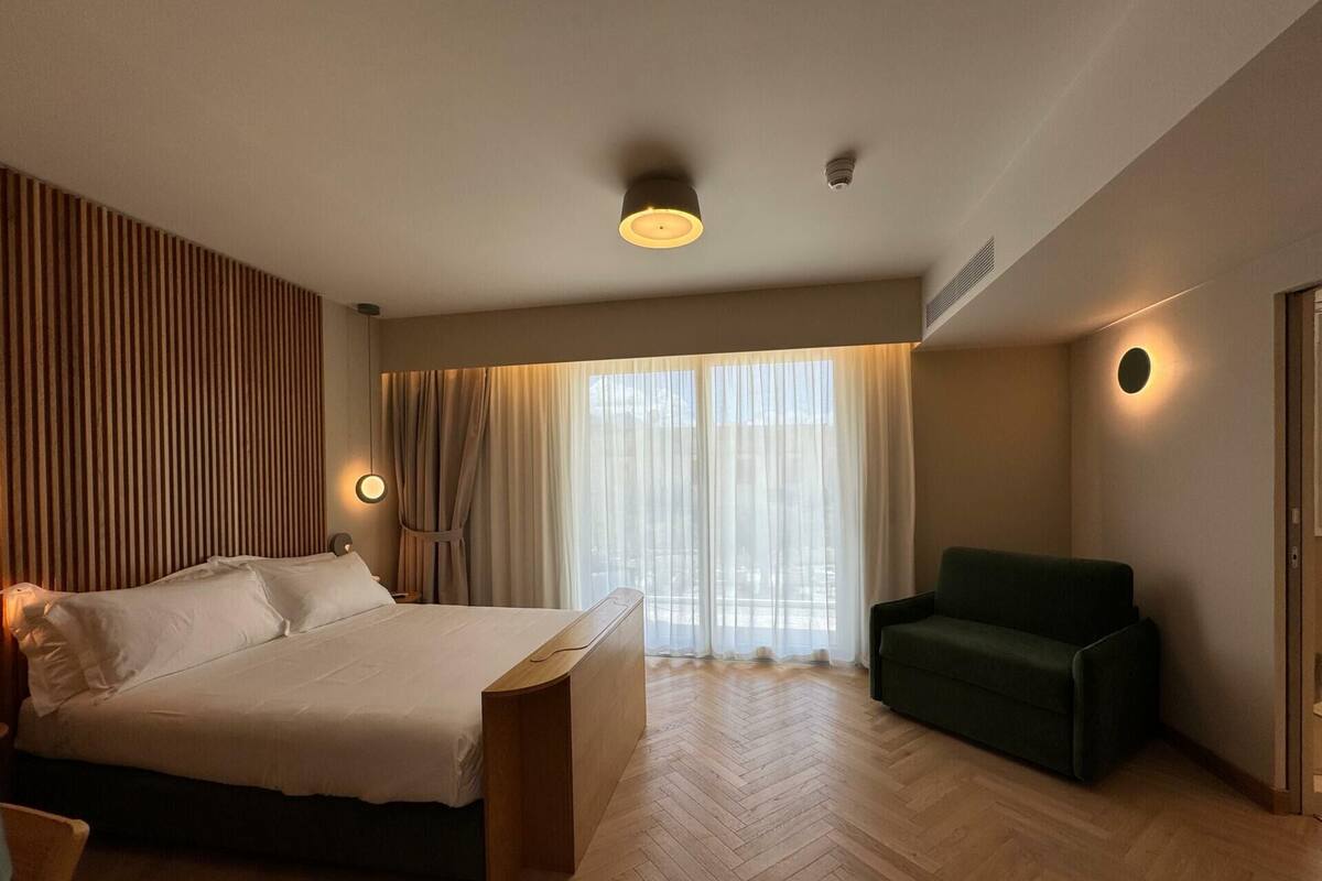 Quarto de hotel com uma cama, uma poltrona verde, piso de madeira e uma janela grande com cortinas.