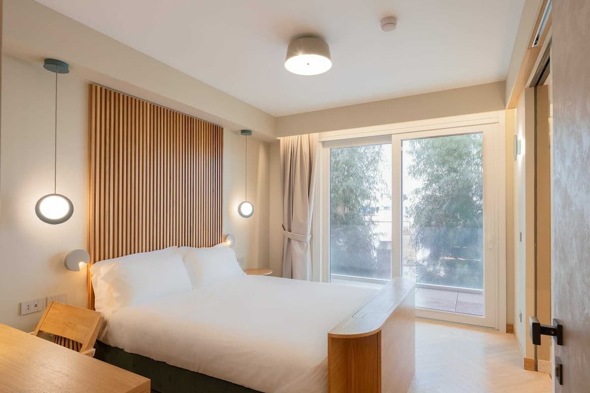 Quarto moderno com uma cama grande, mesas de cabeceira, cabeceira de madeira e uma porta de vidro deslizante que dá para uma varanda.