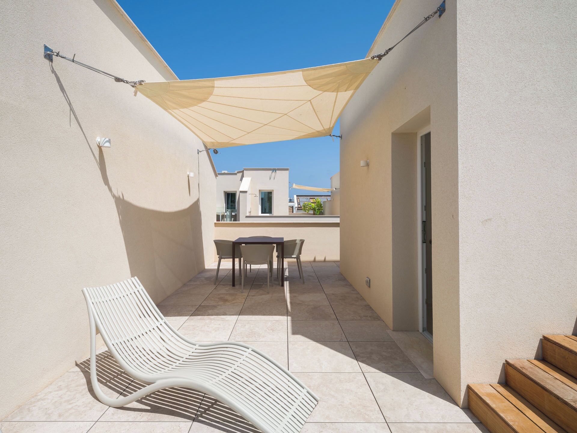 Une terrasse ensoleillée avec une table et des chaises, un fauteuil de jardin blanc et un escalier en bois.
