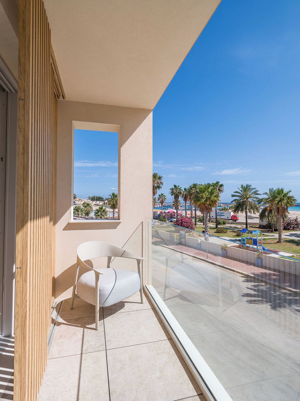 Ein Balkon mit weißem Stuhl, Blick auf Palmen und Strand, und ein klarer Himmel.