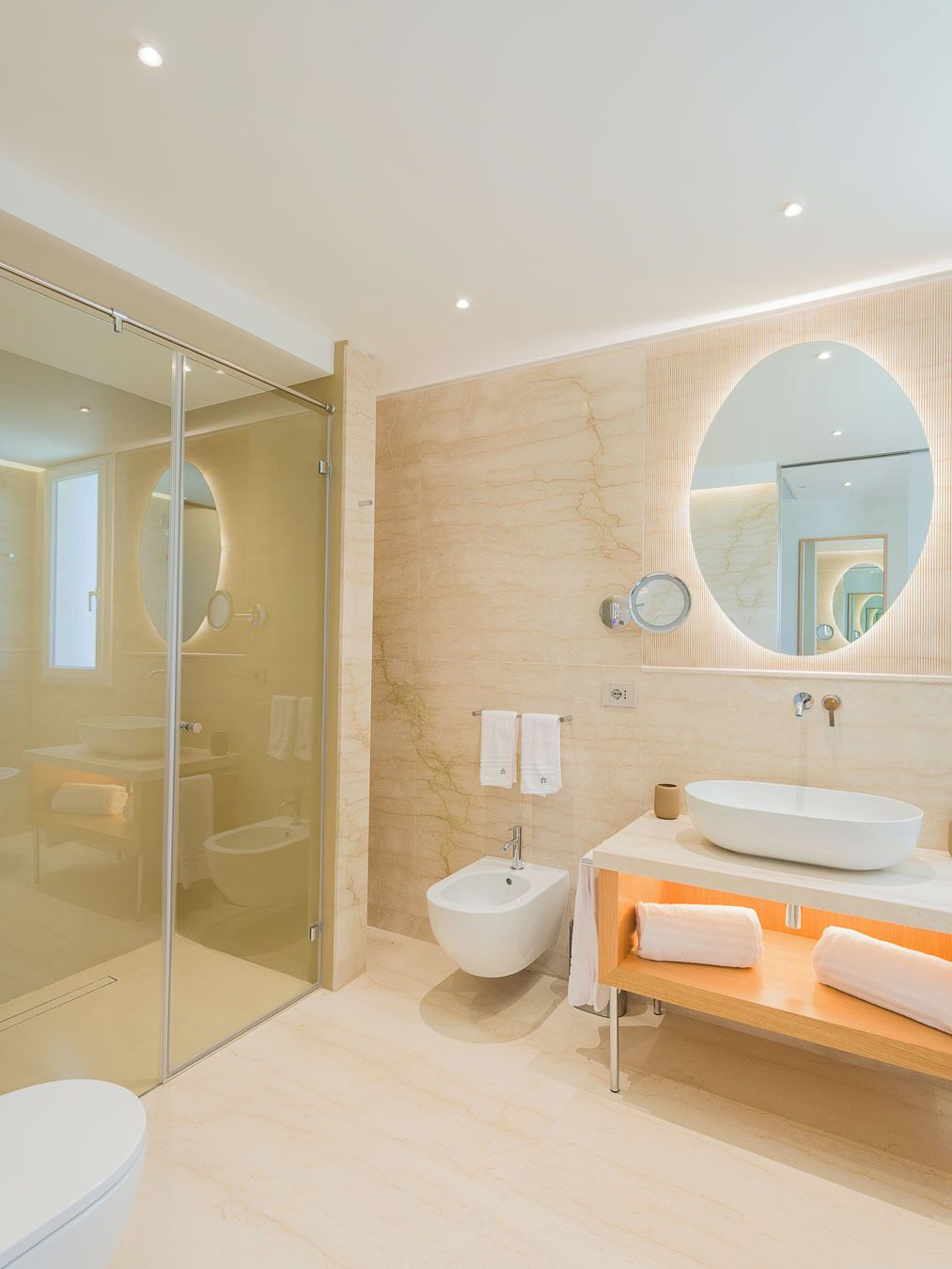 Een moderne badkamer met een glazen douchecabine, een losstaand bidet, een grote ovale spiegel en een houten wastafelmeubel met een wastafel.