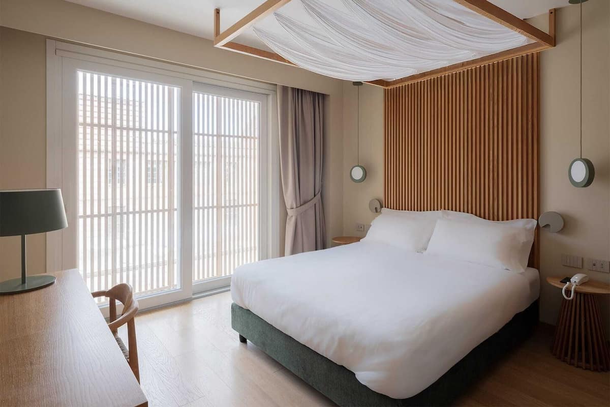 Quarto de hotel moderno com uma cama grande, uma escrivaninha e uma janela com persianas.