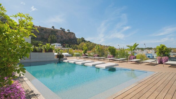 Piscina con sillas de descanso y terraza de madera, rodeada de vegetación y con vistas a una colina.