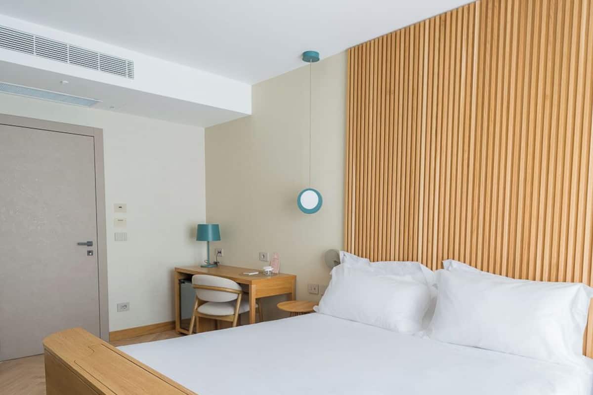 Quarto de hotel moderno com estrutura de cama de madeira, uma escrivaninha com cadeira, uma mesa redonda pequena e uma luminária fixada na parede.
