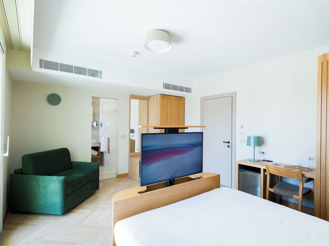 A hotel room with a bed, a TV, a desk with a chair, a green sofa, and a bathroom with a mirror and towel.