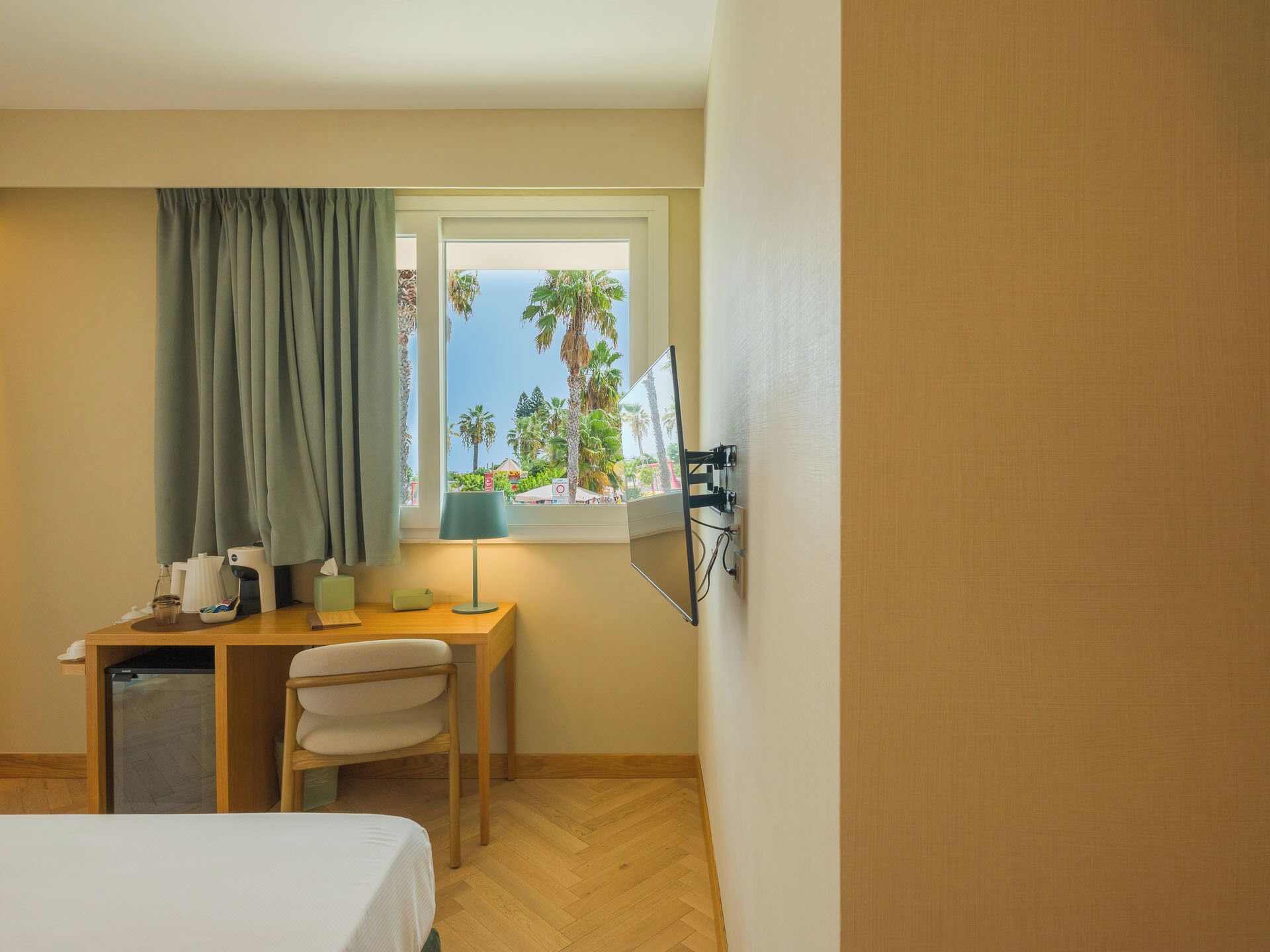 A hotel room with a bed, a desk, a chair, a window with a view of palm trees, and a wall-mounted TV.